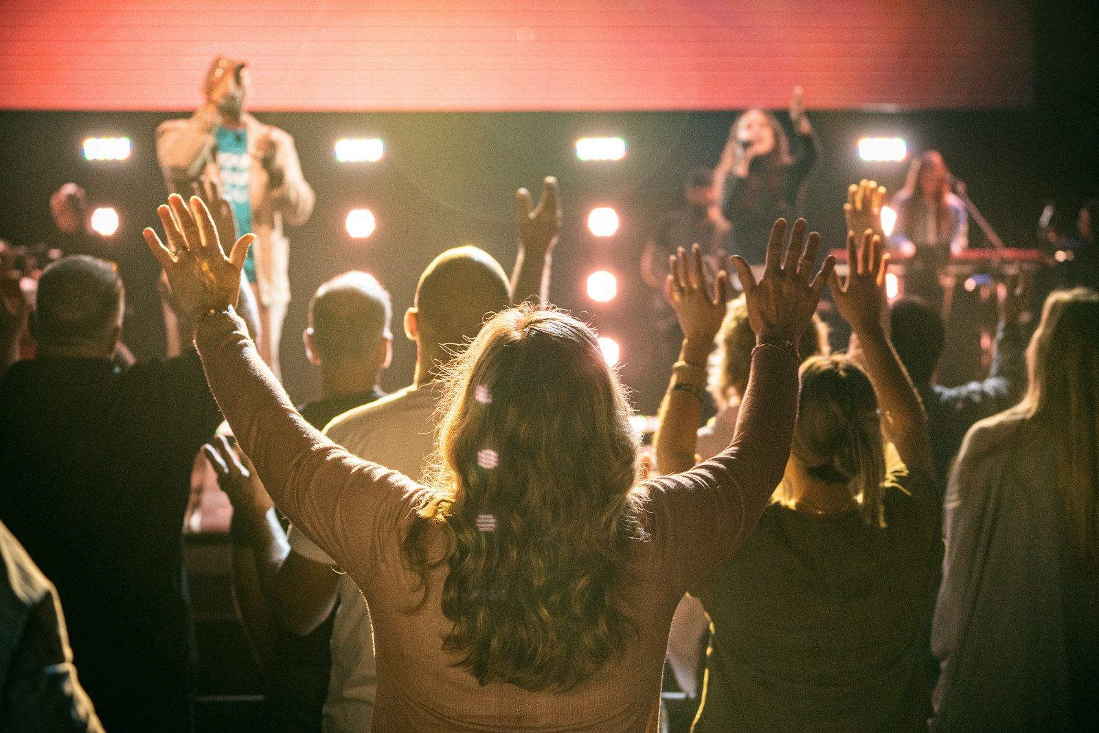 A diverse congregation worshipping together, seen from behind, hands raised toward the stage